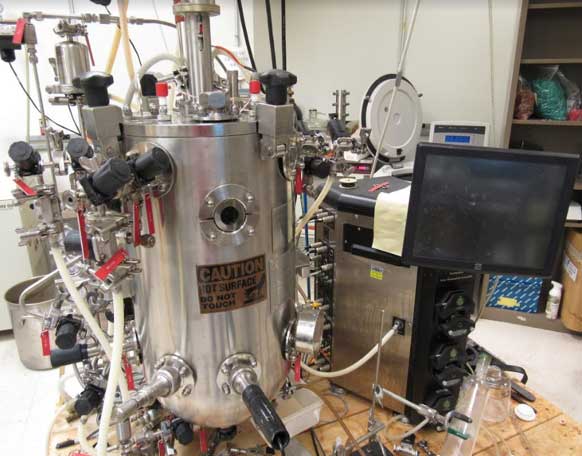 Large stainless-steel bioreactor vessel with attached tubing, control valves, and a caution label reading “Hot Surface Do Not Touch,” positioned beside a monitor and control unit in a laboratory