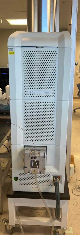 Tall white analytical instrument on wheels with tubing connections, part of a mass spectrometry system in a laboratory