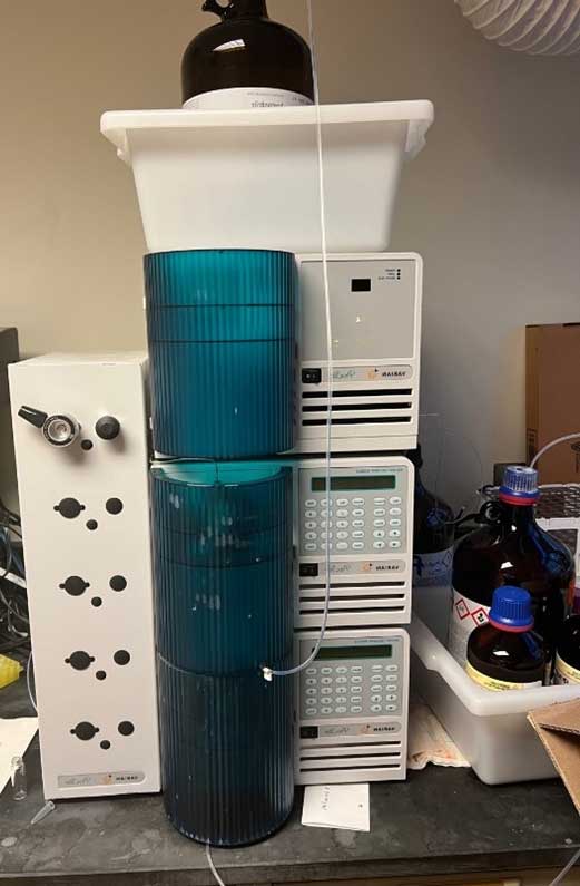 Stacked H.P.L.C. instrument with teal cylindrical components and solvent bottles beside it on a lab bench, part of an Akai system