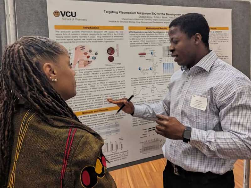 A man gestures while explaining a research poster to a woman at a V.C.U. School of Pharmacy event. The poster, titled “Targeting Plasmodium falciparum Sir2 for the Development of Antimalarial Drugs,” includes text, charts, and diagrams. Both individuals appear engaged in discussion in a professional indoor setting.
