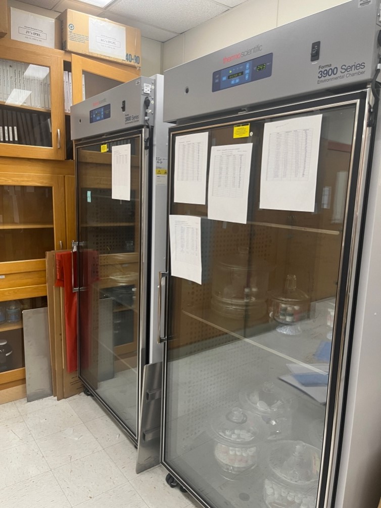 Environmental chambers in a lab workspace with glass doors and posted inventory sheets