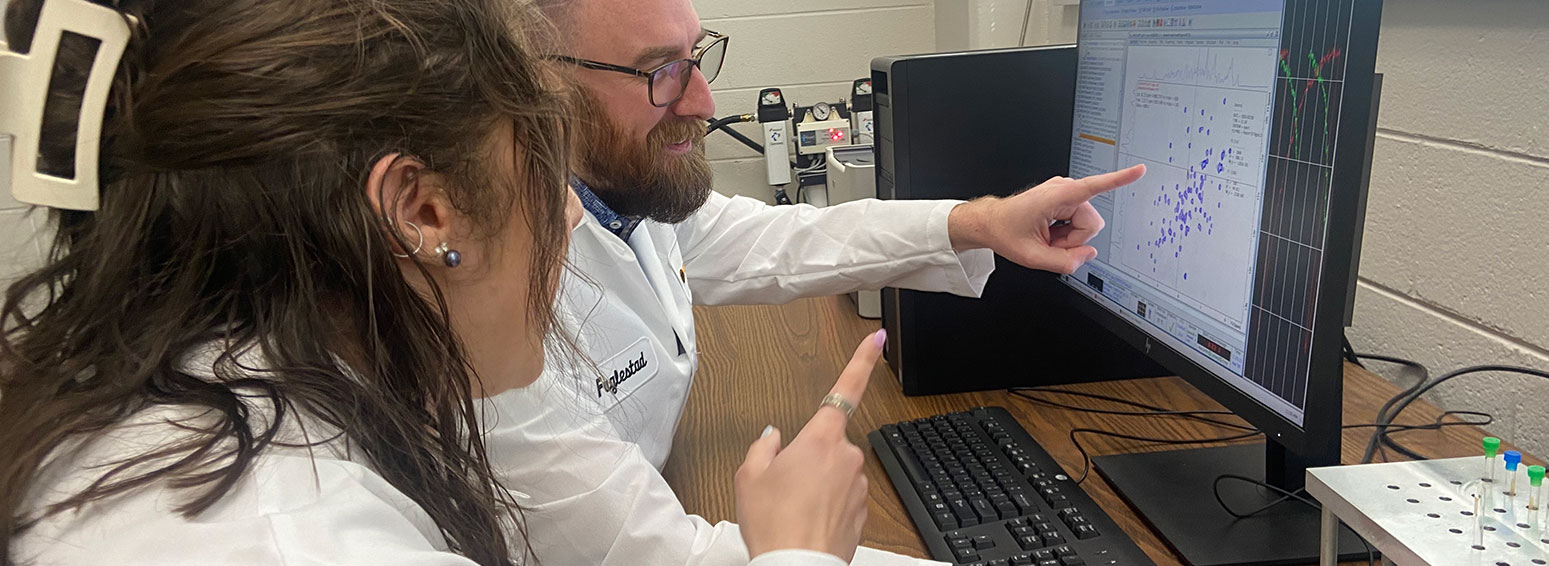 two people are seated at a computer workstation wearing lab coats - they are both pointing to a scatter plot graph on the computer's monitor