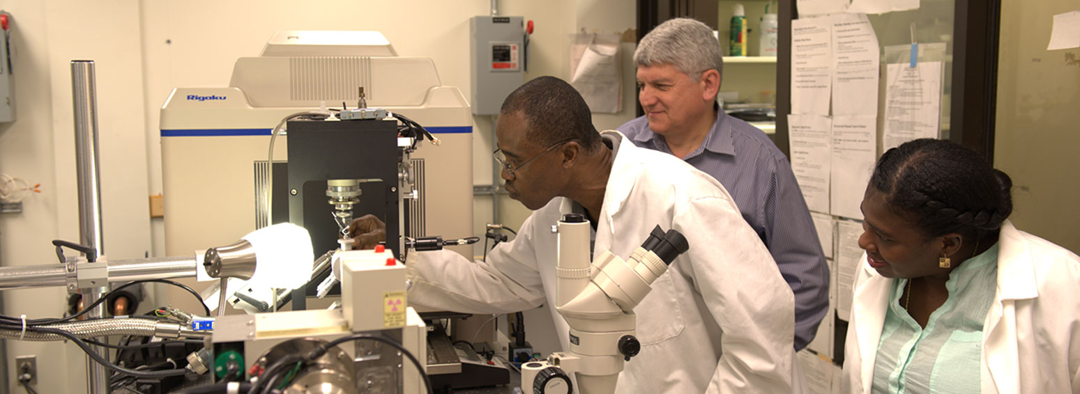 one researcher in a lab coat works intently with a piece of lab equipment while two other smiling people observe closely
