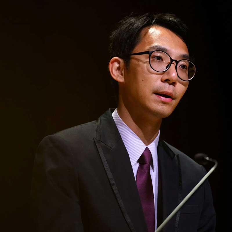 A man in a coat and tie and glass speaks into a microphone.