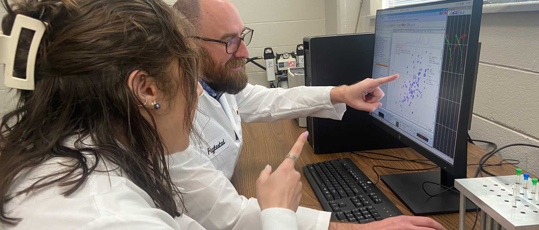 A faculty researcher points to data on a computer screen while discussing results with a student in the lab