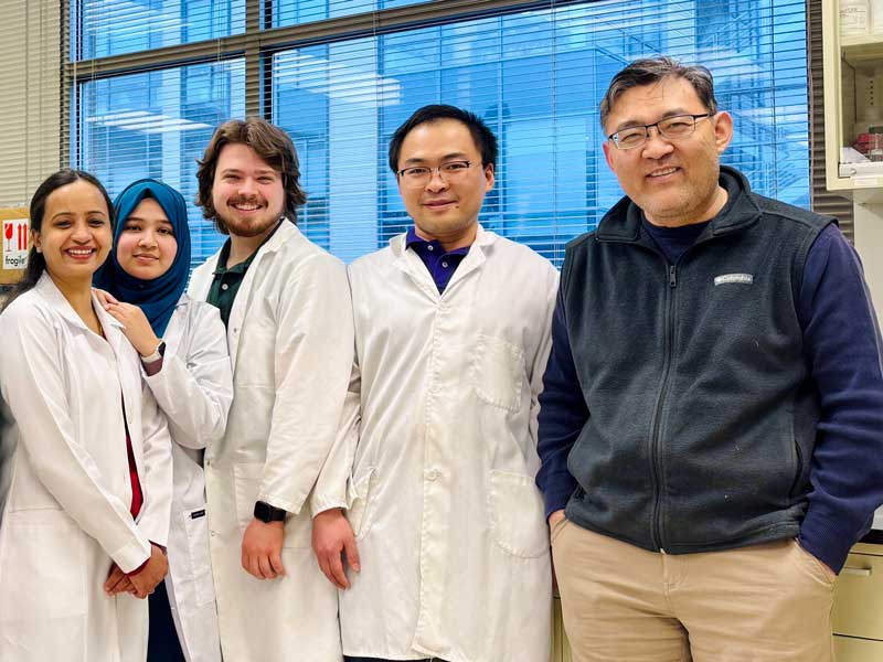 A group of five researchers in lab coats stands together smiling in a laboratory with large windows in the background