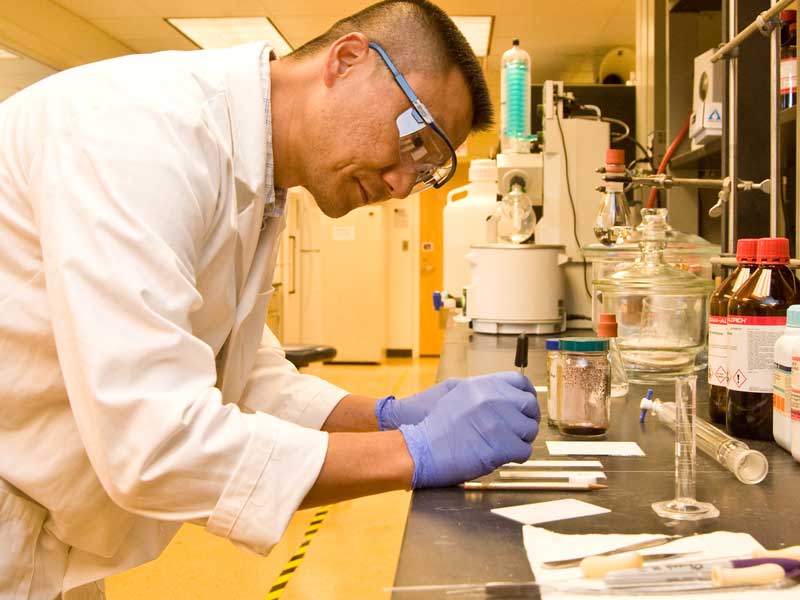 A researcher wearing safety glasses, gloves, and a lab coat conducts an experiment at a laboratory bench with glassware and chemical bottles