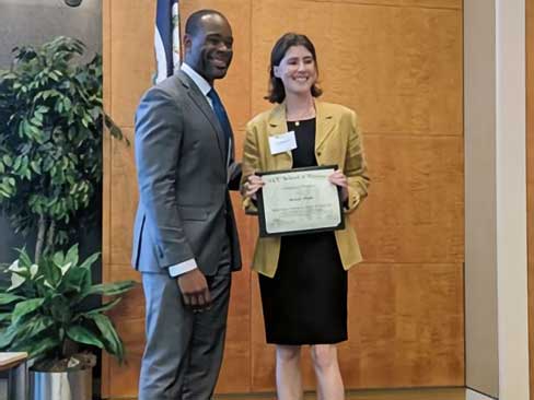 A woman stands beside a man in a gray suit while holding a certificate at a V.C.U. School of Pharmacy event. Both are smiling in a formal indoor setting with a wooden wall, flag, and potted plant in the background.