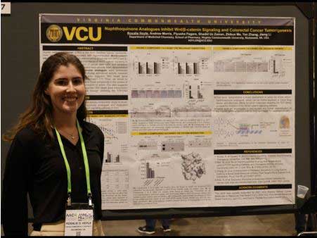 A woman stands smiling beside a research poster titled “Naphthoisoquinone Analogues Inhibit Wnt/β-catenin Signaling and Colorectal Cancer Tumorigenesis” at an event. The poster includes graphs, charts, molecular diagrams, and text detailing the study’s findings. The setting appears to be an academic conference or research symposium.