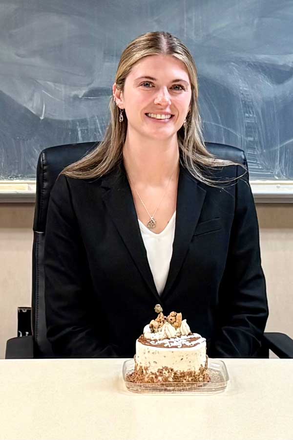 Ally Thompson smiles for a photo with a celebratory cake sitting in front of her on a desk after her dissertation defense.